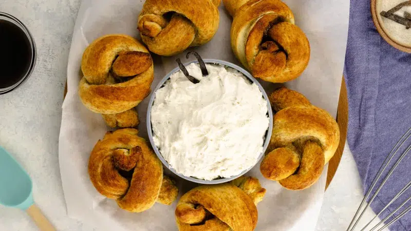 top view of six vanilla bean pretzels surrounding a cream cheese dip with vanilla beans sticking out of dip. The pretzels and dip lay on top of a round dish with white parchment paper. Surrounding the pretzels is a wooden spatula, a clear pink bowl with Rodelle's Vanilla Paste, a silver whisk, blue towel, and spent beans with sugar mixed in with them.