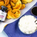 top photo of a clear bowl containing a cream cheese dip laying on top of a bold blue towel. In a smaller clear bowl to the upper right is vanilla bean paste. To the upper left is a pile of vanilla bean pretzels laying on top of white parchment paper and Rodelle's vanilla paste laying on top of