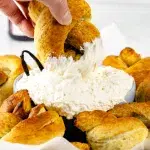 Vertical side profile photo of a hand dipping a vanilla bean pretzel into a large clear glass of cream cheese dip with vanilla beans sticking out on side. Surrounding the dip are more pretzels on a wooden plate with white parchment paper.