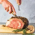 Side profile of a prime rib roast being cut with a carving set by masculine hands. The prime rib lays on top of a medium sized cuttong board with herbs on the side for accent.