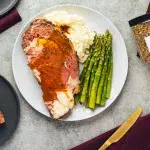 Top view of a white plated prime rib roast with fresh asparagus, mashed potatoes and gravy poured on prime rib and potatoes. Surrounding the plate are charcoal color plates with more prime rib, Rodelle's Prime Rib seasoning, brass eating utensils, and maroon colored cloth napkins.