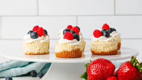 side profile of multiple mini vanilla bean cheesecakes on top of a white cake stand. In the background is white tile, a light turquoise blue towel, and in the foreground is a group of large strawberries. Topping the mini cheesecakes is a swirl of whipped cream, raspberries and blueberries.