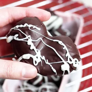 Close up of a hand holding a chocolate covered strawberry heart with white chocolate drizzle