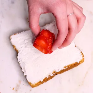 Hand placing strawberry flower cut out on brioche bread with whipped cream on top