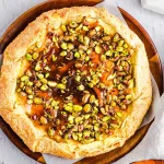 Galette made with flakey crust, sweet cheese filling and pistachios topped on a wooden serving dish and on a neutral background