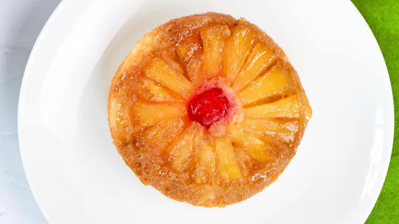 top close up shot of pineapple upside down cake sitting on a white plate with a lime green colored towel next to plate.