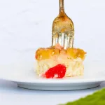 side view of piece of pineapple upside down cake with antique rustic fork puncturing it from the top. A bit of maraschino cherry is seen in front of the piece of cake.