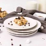 no bake rocky mountain cookies laying on top of a stack of white plates. Gray towel lays in the background and a large plate of more cookies. Marshmallows and chocolate chip cookies sprinkle around the scene.