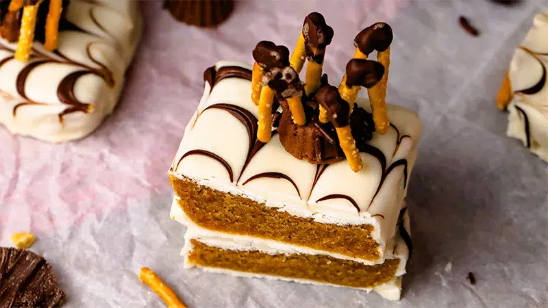 Top angle view of a peanut butter candy bars that's been cut in half, each piece on top of one another. The bar is covered in white chocolate with a spider web pattern made with dark chocolate. A homemade pretzel Reese's spider lays on top of the bar. The bar is surrounded by parchment paper and other halloween themed bars.