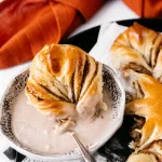 a piece of the star bread laying in the white glass bowl of browned butter vanilla bean glaze. Surrounding the star bread piece is the rest of the star bread on a black plate and an autumn red towel.