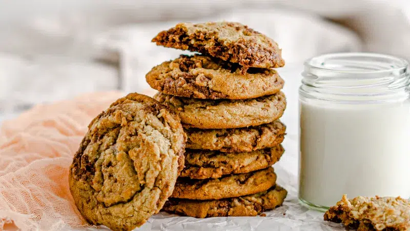 Side shot of a stack of seven halva cookies, one cookie is propped up next to the stack of cookies. Surrounding the cookies is a pink scarf, and a glass of milk.