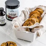 Angled shot of a group of halva cookies in a bread tin with a light gray towel. Laying on the side is Rodelle's Gourmet Baking Cocoa Canister and Rodelle Pure Madagascar Bourbon Vanilla Extract.