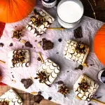 Top view of a bunch of homemade spider web chocolate peanut butter protein candy bars. Pretzels and chocolate are scattered around the scene along with some medium to small pumpkins and glass of milk.