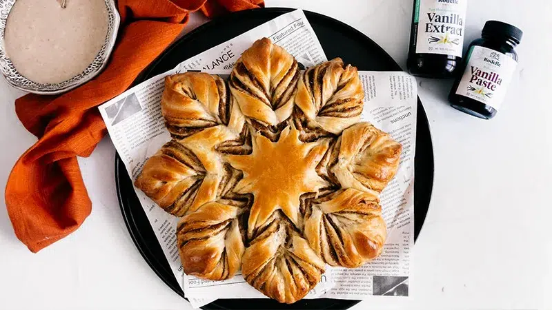 Top shot of Fig Jam Star bread on parchment paper and a black plate. To the right is Rodelle's Pure Madagascar Bourbon Vanilla Extract and Rodelle Natural Vanilla Paste. To the left is an autumn red towel and the Browned Butter Vanlla Bean Glaze in a bowl.