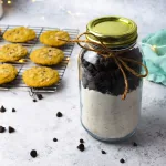 an angled shot of a 16oz jar containing a diy cookie mix with a top layer of chocolate ship cookies. The mason jar has a gold lid on top and a bow made out of twin wrapping the top portion for decorations. behind the jar is a light blue towel and to the left is a cooling rack with chocolate chip cookies. Chocolate chips surround the the photo and are scattered.