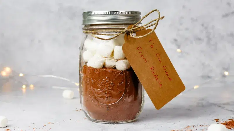 Side profile of a homemade diy hot cocoa mix in a jar with marshmallows. Twine holds a kraft paper take that reads 