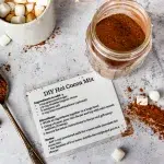 top shot of an opened jar of diy hot cocoa mix and a hand made recipe card for the hot cocoa mix. Surrounding both are scattered marshmallows and hot cocoa mix on the counter top. A spoon hold a bit large amount of the hot cocoa mix. Upper left is a white mug with hot cocoa and marshmallows.