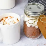 Side angle view of diy hot cocoa mix with a kraft holiday tag held by twine. Next the jar is a white mug with hot cocoa and marshallows on top. In the background is a grey and white striped cloth and white jars.