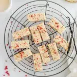 Top shot of pink peppercorn shorbread cookie slices on a black wired round cooling rack. Surrounding the image are bits of pink peppercorn, 2 vanilla beans, small white bowl, and a white cloth.