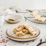 Portrait side image of pink peppercorn shortbread laying on a white with brown speckled plate. Surrounding the shortbread are pink peppercorn flakes, 2 vanilla beans, white bowl, and a cooling wrack with shorbread.