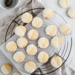 Top view of vanilla peppermint shortbread with white chocolate laying on parchment paper and a black wire cooling rack. A gray kitchen towel and bowl with spoon are in the shot as well.