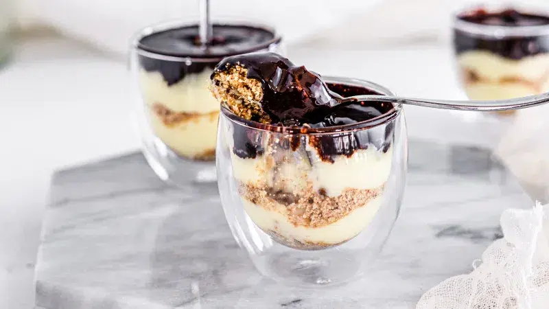 close up of eclair cake parfait. Layers of eclair cream, graham cracker crust, and a top layer of chocolate syrup. A spoon with some of the eclair cake parfait sits on top of the clear glass parfait dish. Parfait stands on a marble decorative board and white background.