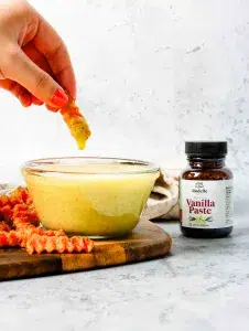 Vanilla aioli sauce in a clear glass container. A hand has dipped a sweet potato fry into it and pulling up showing the drip of aioli sauce. On the side is Rodelle's vanilla paste