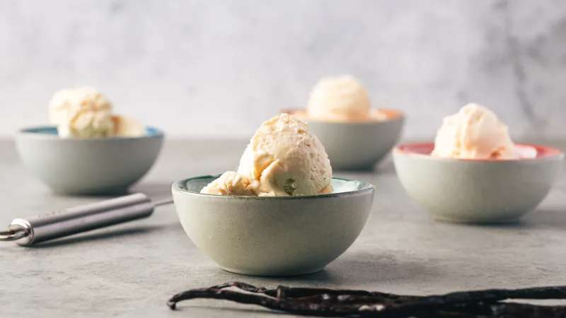 Close up of scoops of no churn vanilla ice cream in small ice cream bowls with a blue-ish green exterior but color interior including, green, pink, orange, red. There is one ice cream bowl up front and three in the background. A pile of vanilla beans are at the bottom right and a silver ice cream scoop in the background. The back drop and table are gray.