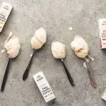 Scoops of ice cream laying in four different sized vintage spoons on a gray backdrop. In the left corner is a white towel with blue stripe and all four 1oz single origin vanilla extracts scattered throughout the scene which includes Uganda, Madagascar Bourbon, Papua New Guinea, and Tanzania.