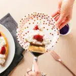 top shot of a slice of an air fryer vanilla cake on a silver pie server being placed on a festive pink foiled with white background paper plate. A hand is gently putting the slice on the plate which is being held by another hand. In the background is the larger portion of the vanilla air fryer cake with whipped cream and sliced strawberries on a white plate. Underneath the white plate is a gray cloth napkin. To the bottom right is a pink satin fork.