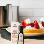 side image of rodelle's Ugandan pure vanilla extract - both the dark amber bottle and box. In the background is the air fryer vanilla cake topped with sliced strawberries and whipped cream, PLUS the air fryer try. The background is white tile on pink table.