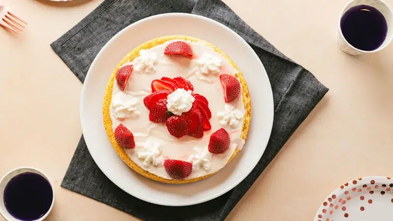 Top photo of an air fryer vanilla cake topped with a vanilla glaze, whipped cream, and fresh strawberries cut in half.