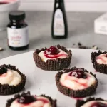 Close up of a cherry cordial tartlet with oreo crumble tart, vanilla bean custard, and a single cherry. In the background are rodelle's papua new guinea pure vanilla extract, and rodelle paste on a gray textured background.