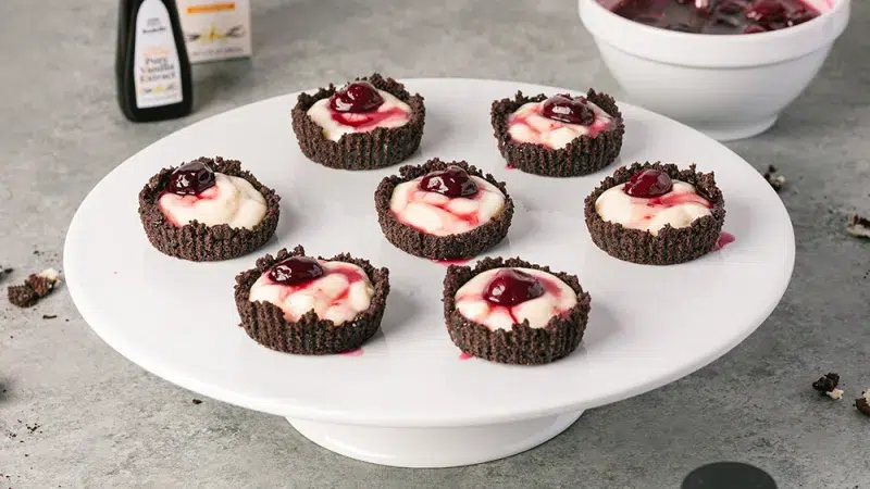 close up of seven cherry cordial tartlets on a white cake stand. In the background is Rodelle's bottle in a box, papua new guinea vanilla extract and a white bowl containing cherry syrup. On the gray textured table are crumbled pieces of oreo.