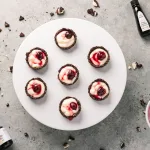 Top view of seven cherry cordial tartlets laying on top of a large white cake stand. Surrounding the tartlets are oreo crumbs, rodelle's 2oz vanilla paste, 2oz papua new guinea vanilla extract, and a white bowl with cherry sauce.