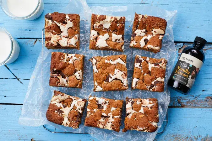 S'mores treat bars cut on parchment paper on a blue wooden background with a cup of milk and bottle of vanilla extract