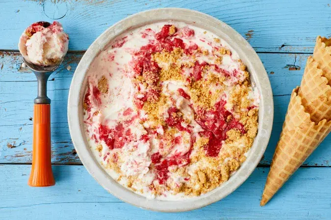 Giant bowl of strawberry cheesecake ice cream on blue wooden background with ice cream scoop and cones in frame