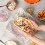 An angled shot of a hand holding the assembled greek chicken gryos. Surrounding the assembled gyro and hand are all ingredients to make the gyros including greek seasoned chicken on an orange plate, pita bread on a yellow plate, homemade tzatziki sauce in a pinch bowl with spoon, crumbled feta in a clear bowl, red onion in a clear bowl, cherry tomatoes cut in half, and a bottle of rodelle's greek seasoning.