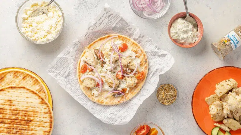 Top view of a plate with greek chicken gyros assembled. On a large pita bread are tomatoes, greek seasoned chicken, onion, feta, and a homemade tzatziki sauce. Surrounding the gyro which is on some parchment paper are all the ingredients to assemble the gryo. The background is white.