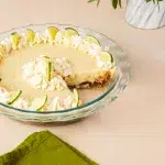 Angled shot showing a slice cut out of the key lime pie to show a creamy yummy pie filling. The key lime pie is in a clear glass pie dish. In the background is a green plant in a white and gray marbled pot. In the foreground is a green towel and piping bag.