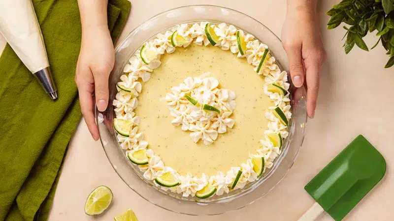 Photo taken from above of a whole key lime pie with flecks of vanilla seeds in the pie and a star like frosting pattern on the top with slivers of lime. Hands are on both sides of the clear pie dish holding the pie. Surrounding the pie is a green spatula, green towel, frosting piping bag, and a green plant. The background in a like orange pink color.