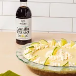 side angle view of a vanilla key lime pie in a clear glass pie dish. The pie has a graham cracker crust and key lime pie filling with flecks of vanilla seeds and lime. On top of the pie is a star frosting pattern with triangular lime slices sticking out. In the background is Rodelle's 16oz Pure Madagascar Bourbon vanilla extract and a white tiled background. The table is a light pink, orange color. In the foreground is a green cloth and a piping bag with frosting.