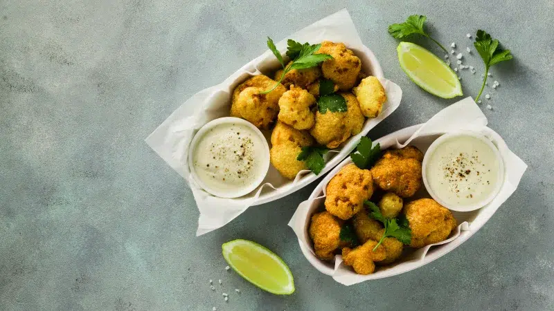 Top view of two white flat oval bowls with parchment paper containing Sriracha Honey Cauliflower Wings and a small white glass dish containing homemade lime greek yogurt dip. The two servings are on the right side of the image with two sliced limes on both sides and cilantro sprinkled throughout the scene. The background is a gray marble.