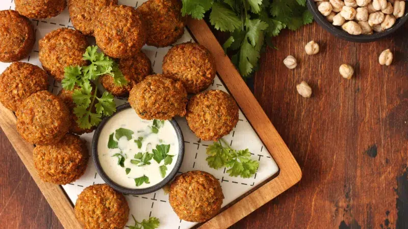 A group of chickpea fritters on a wooden platter with white accent. In the group of fritters is homemade tzatziki sauce. Parsley is used as garnish on fritters and in sauce. The background is a medium wood with chickpeas in a charcoal colored bowl.