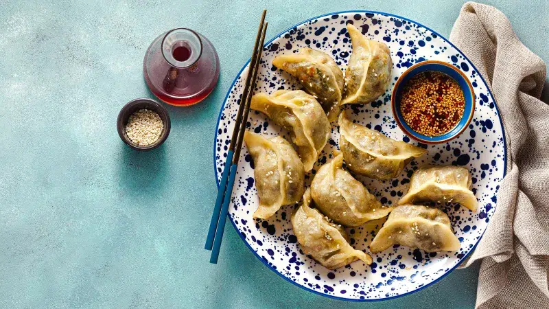 Above shot of Shrimp Potstickers with Soy Chili Sauce. Eight pot stickers lay on a blue and white patterned plate on side are black and blue chop sticks. To the right of the plate is a neutral gray towel. The counter is turquoise.