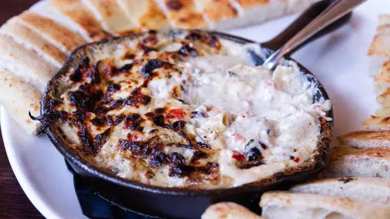 close up of a creamy hot cream dip in a small black skillet surrounded by crackers. A portion of the crab dip has been eaten and area not eaten is toasted on top.