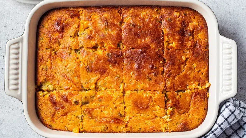 Top view of a 9x12 inch ceramic white pan containing Honey Sriracha Spicy Cornbread. It's cut into 4x3 squares. The background is a light gray and to the right is a blue and white striped dish towel.