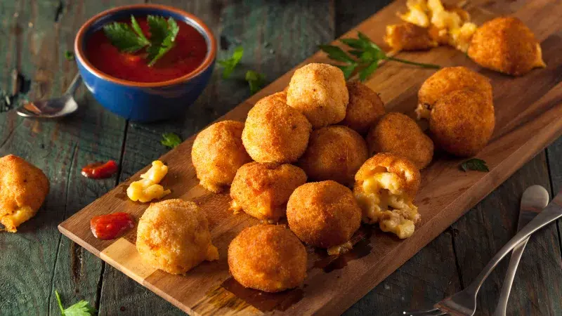 Angle shot of Spicy Mac n Cheese Bites on a wooden platter. In a small darker colored sauce cup is ketchup. The background is dark gray.