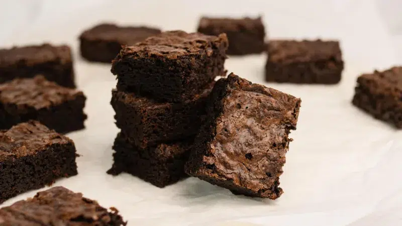 Square cut up gluten free brownies. Three are stacked in the center of the photo and one lays on it. Surrounding the center brownies are more brownies on a white background