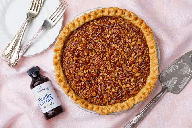 caramel pecan pie on pink tablecloth with plate and serving utensils with bottle of vanilla extract